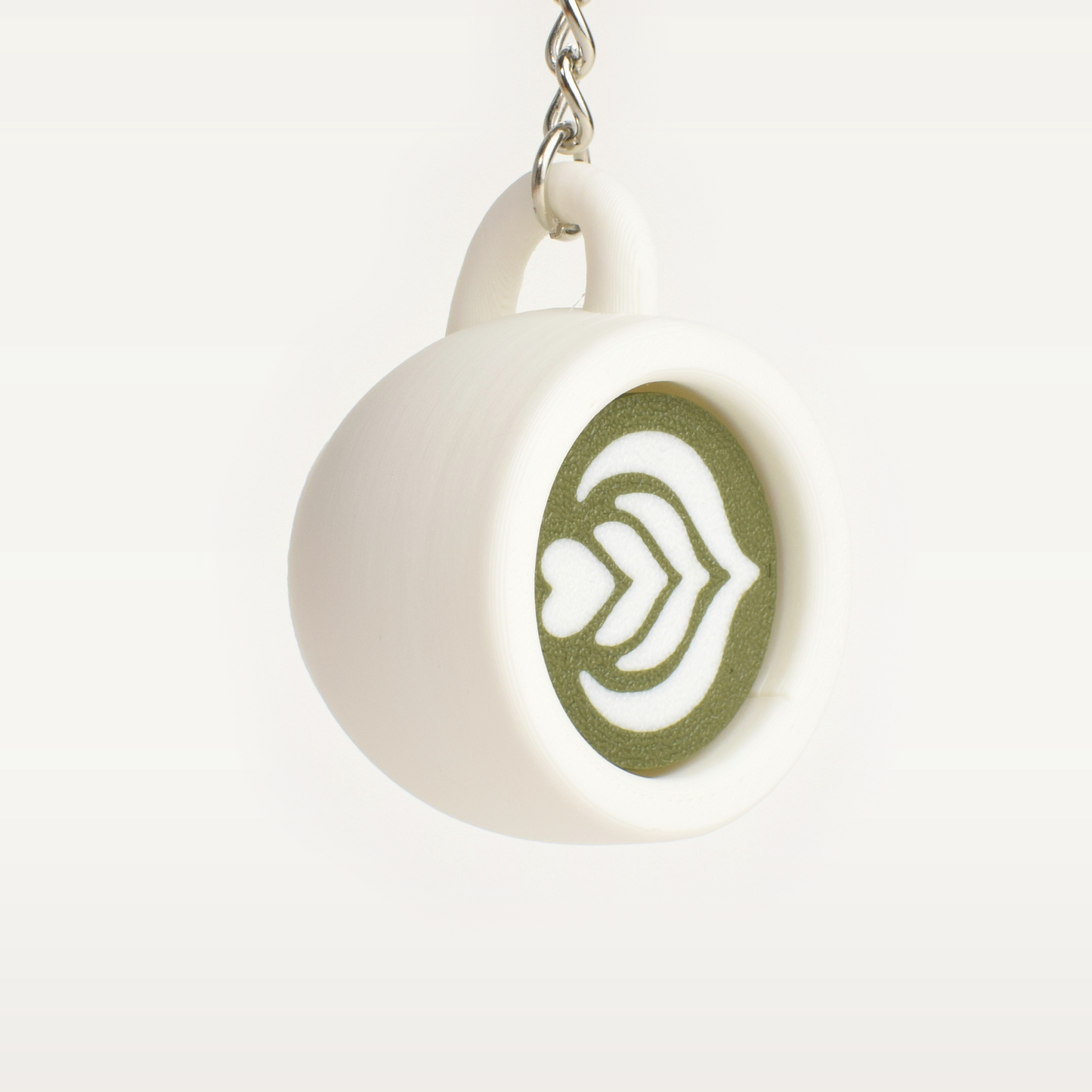 A white ceramic coffee mug keychain with a green matcha latte art on the front, hanging from a silver metal chain against a light gray background.