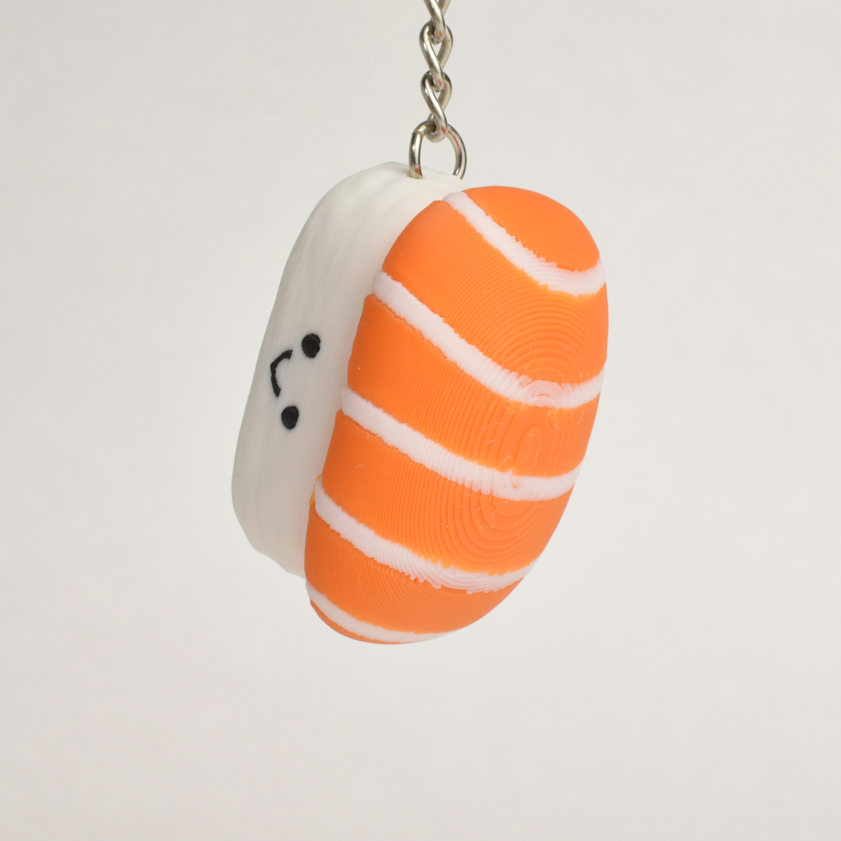 A small sushi-shaped clicker keychain with a smiling face, featuring a salmon top and a white rice base, attached to a silver metal key ring on a plain white background.