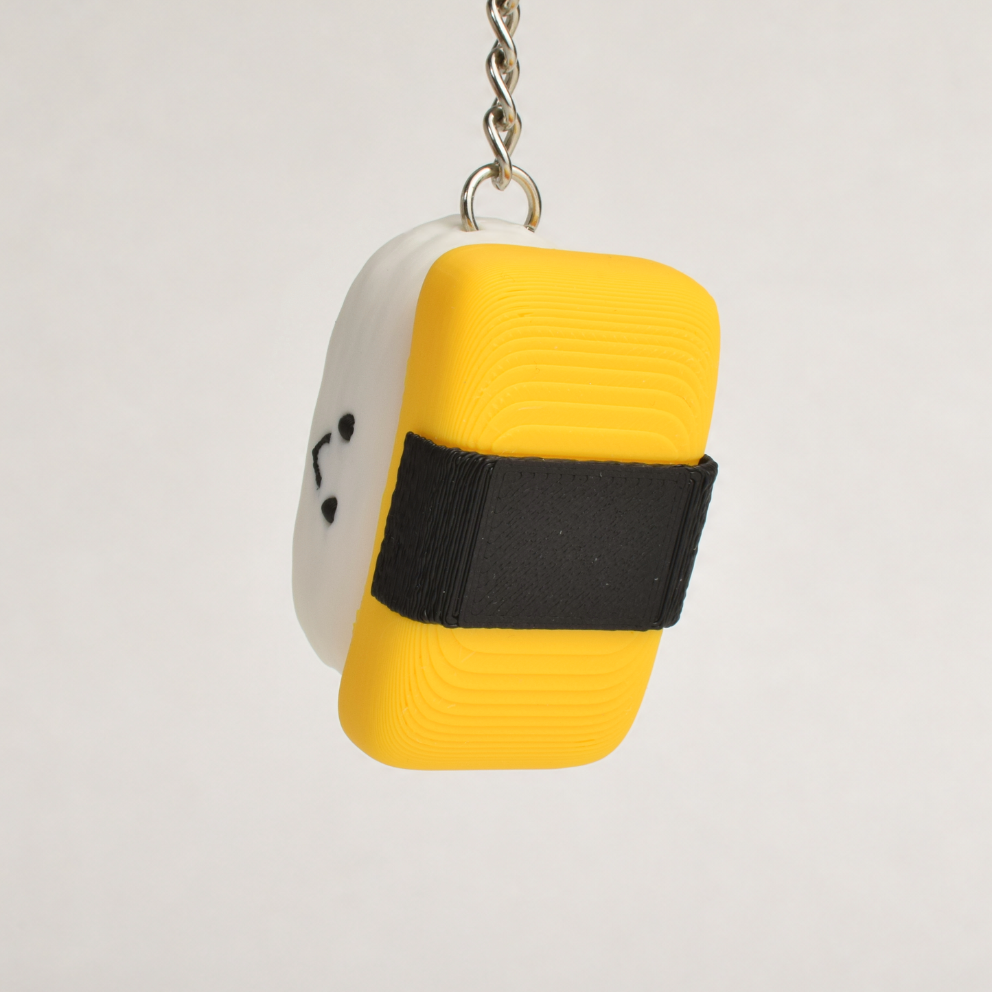 A small sushi-shaped clicker keychain with a smiling face, featuring an egg and seaweed top and a white rice base, attached to a silver metal key ring on a plain white background.
