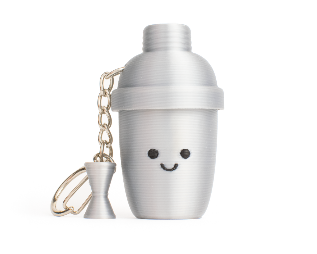 A silver-aluminum-color cocktail shaker fidget clicker keychain with a cute smiling face and a small matching hourglass-shaped measuring cup charm, set against a plain white background.
