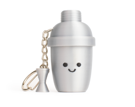 A silver-aluminum-color cocktail shaker fidget clicker keychain with a cute smiling face and a small matching hourglass-shaped measuring cup charm, set against a plain white background.