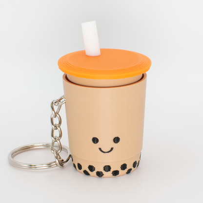 A fidget clicker resembling a cup of bubble tea with a smiling face, an orange lid, and a straw, attached to a keychain.