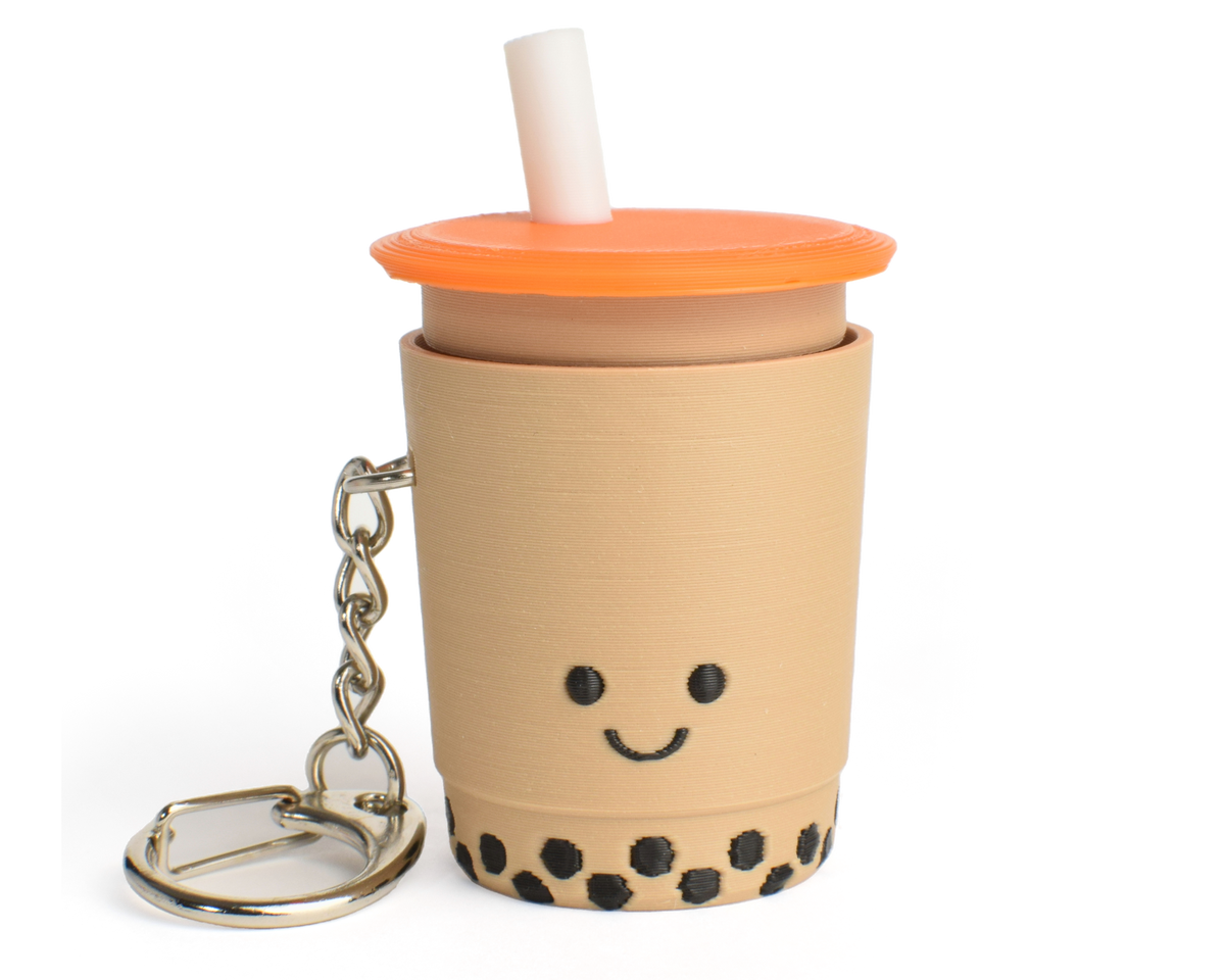 A fidget clicker resembling a cup of bubble tea with a smiling face, an orange lid, and a straw, attached to a keychain.