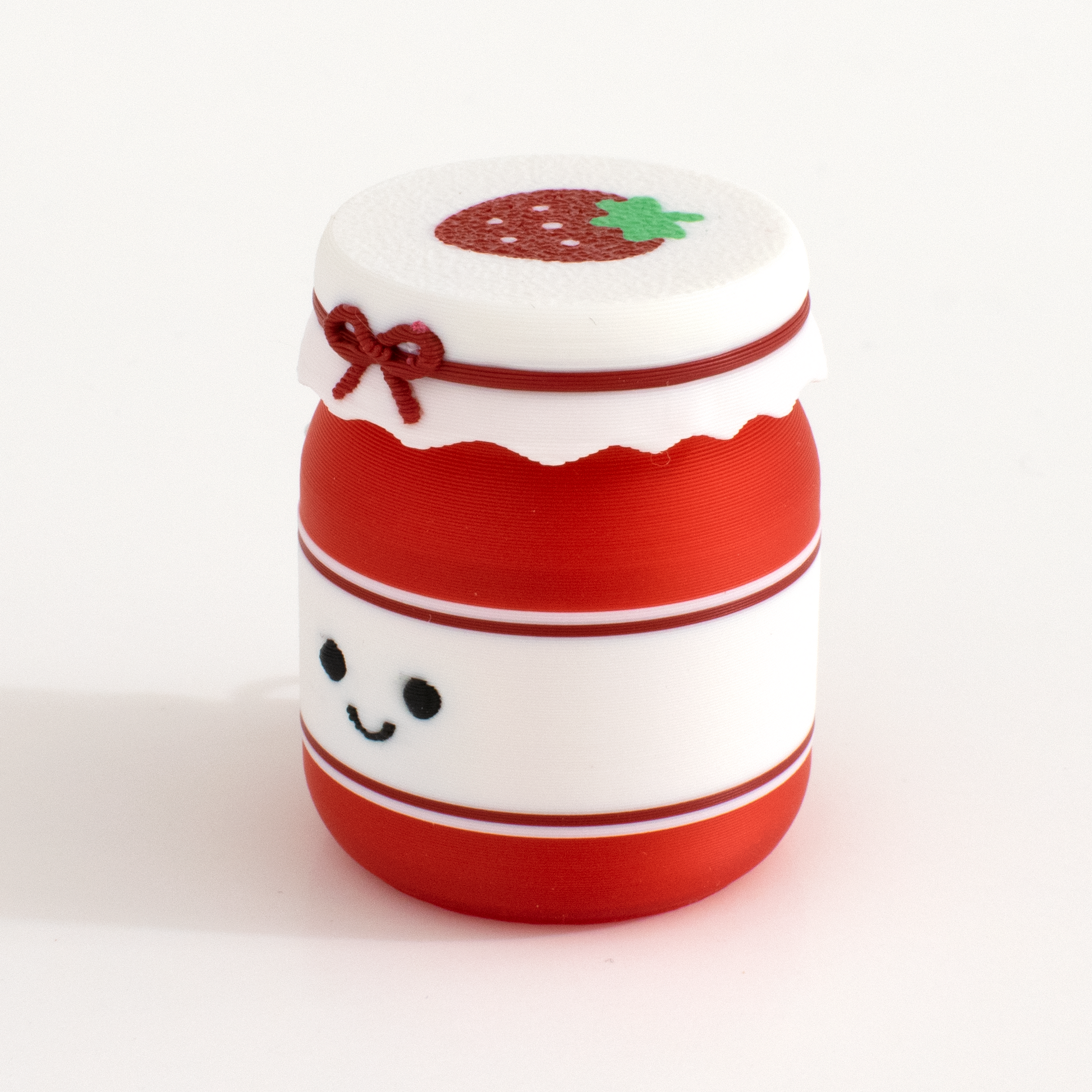 A small strawberry jam mar clicker with a red and white body, a white lid decorated with a strawberry and bow, and a cute black smiley face on the front, photographed on a clean white background.