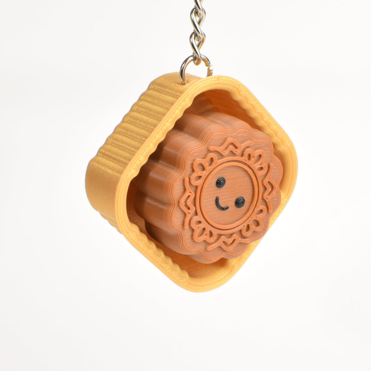 A small hanging charm shaped like a mooncake, with a gold outer wrapper and a brown inner mooncake featuring a simple smiley face, suspended against a plain white background.