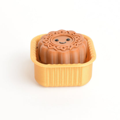 A small brown mooncake-shaped clicker with a smiling face sits inside a gold plastic container on a plain white background.