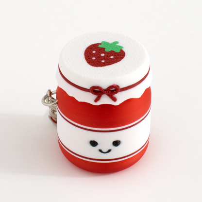 A small strawberry jam jar clicker or bag charm, featuring a white top with a textured strawberry design, a red band with a red bow, a smiling face on the front, and a silver metal key ring.