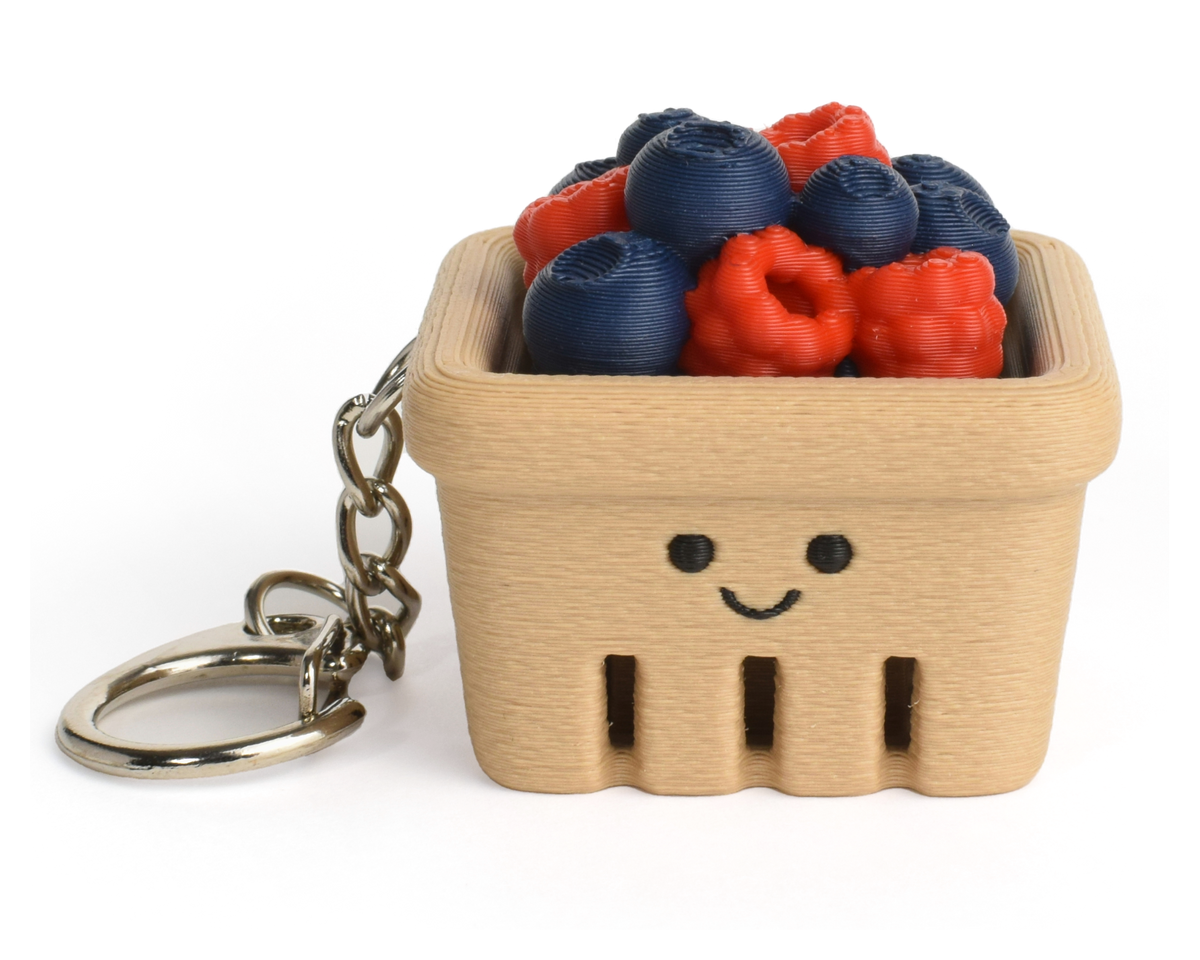 A small beige berry basket keychain with a smiling face, filled with faux blueberries and raspberries, attached to a silver metal key ring on a white background.