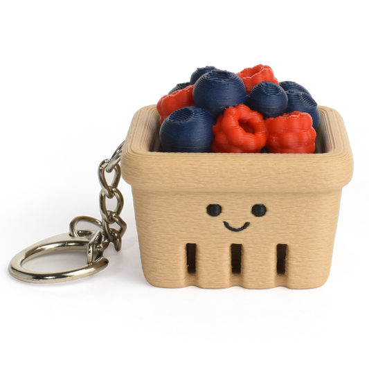 A small beige berry basket keychain with a smiling face, filled with faux blueberries and raspberries, attached to a silver metal key ring on a white background.