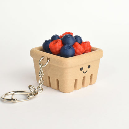 A miniature berry basket keychain with a smiling face, filled with small blueberries and raspberries, attached to a silver metal split ring on a white background.
