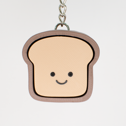 A fidget clicker toy designed to look like a smiling loaf of bread with a beige and brown color scheme, hung on a keychain that could serve as a bag charm