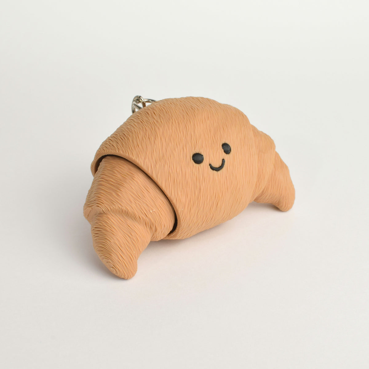 A small croissant-shaped clicker keychain in a warm tan/beige color, with a simple smiley face and a silver metal key ring at the top, photographed on a clean white background.