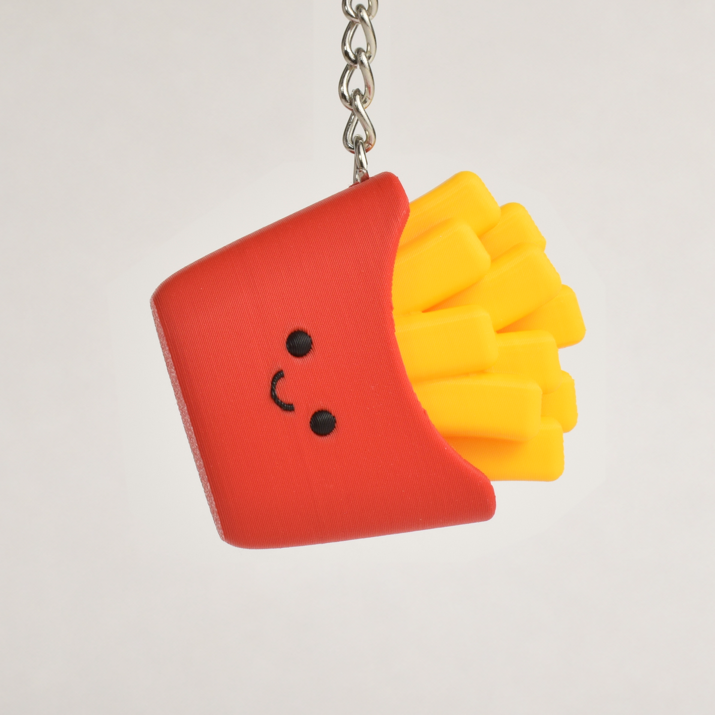 A small novelty keychain shaped like a smiling french fries container, with red packaging and yellow fries, hanging from a silver chain against a plain light background.