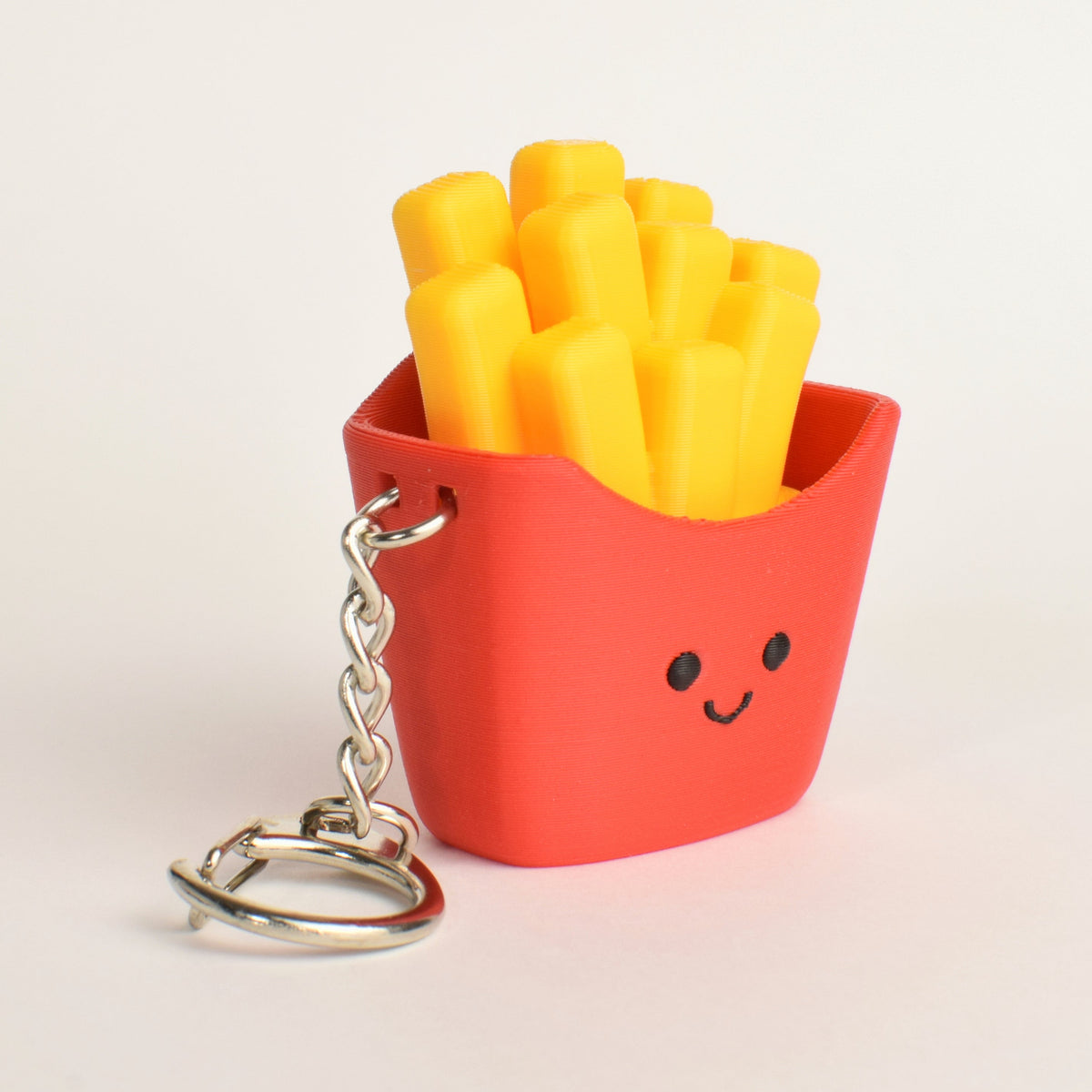 A cute, small keychain shaped like a red french fries container filled with yellow fries, featuring a smiling face and a silver metal keyring on a clean white background.