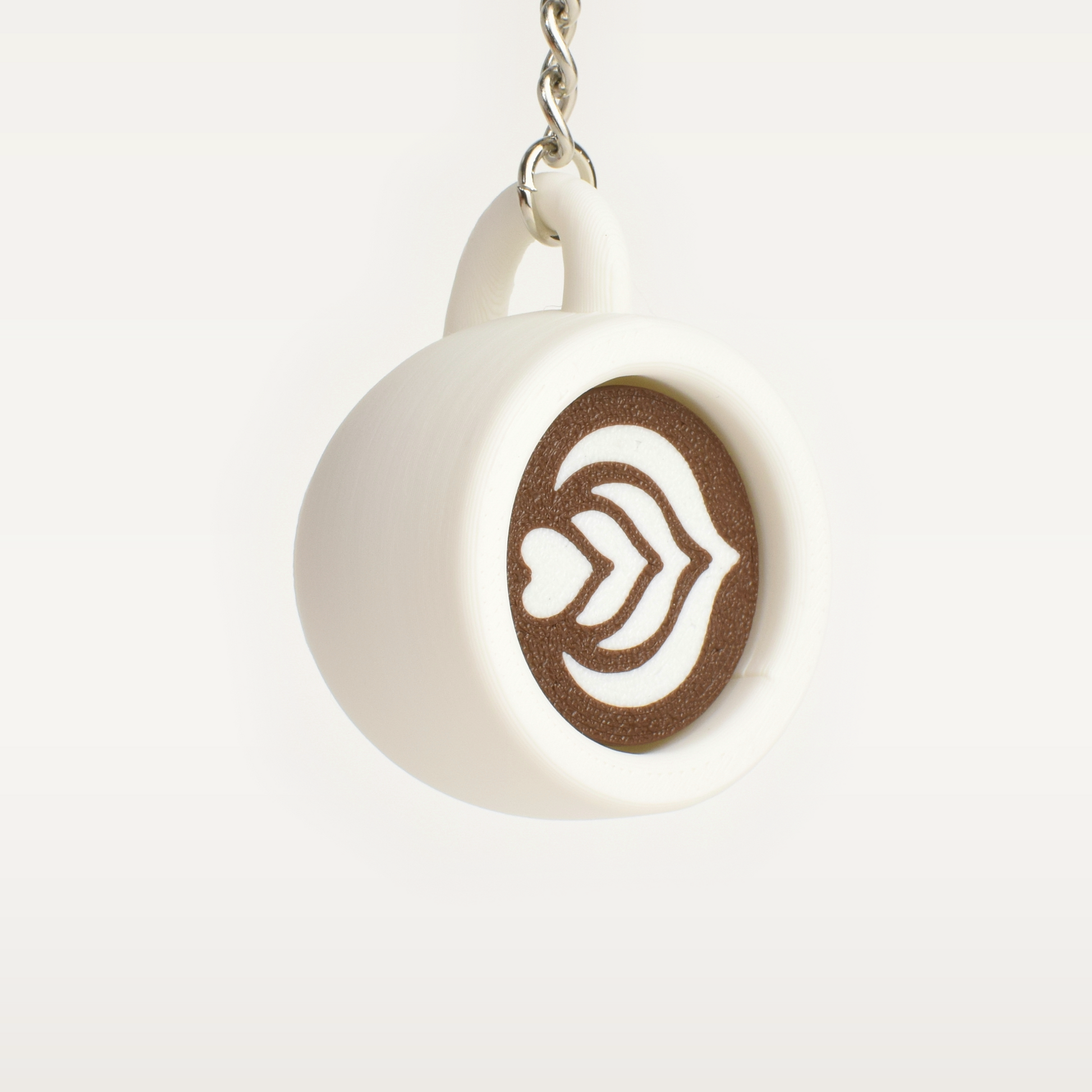 A small white ceramic coffee mug clicker keychain with brown coffee latte art, hanging from a silver metal chain against a light off-white background.