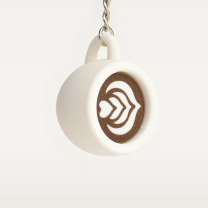 A small white ceramic coffee mug clicker keychain with brown coffee latte art, hanging from a silver metal chain against a light off-white background.