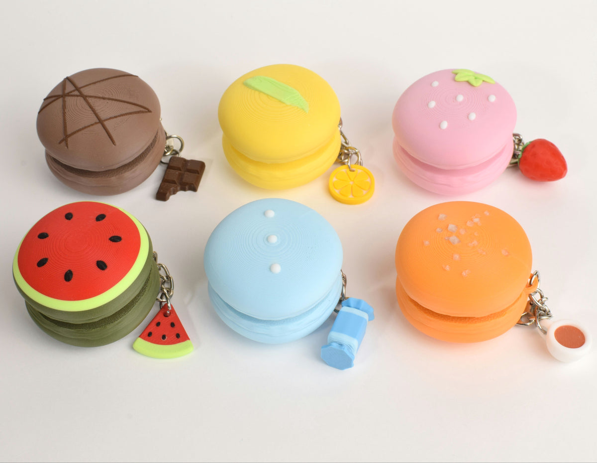 Six cute macaron clickers  in assorted colors, including chocolate (brown), lemon(yellow), strawberry(pink), watermelon(red and green), chai(orange) and bubblegum(light blue), arranged on a clean white background