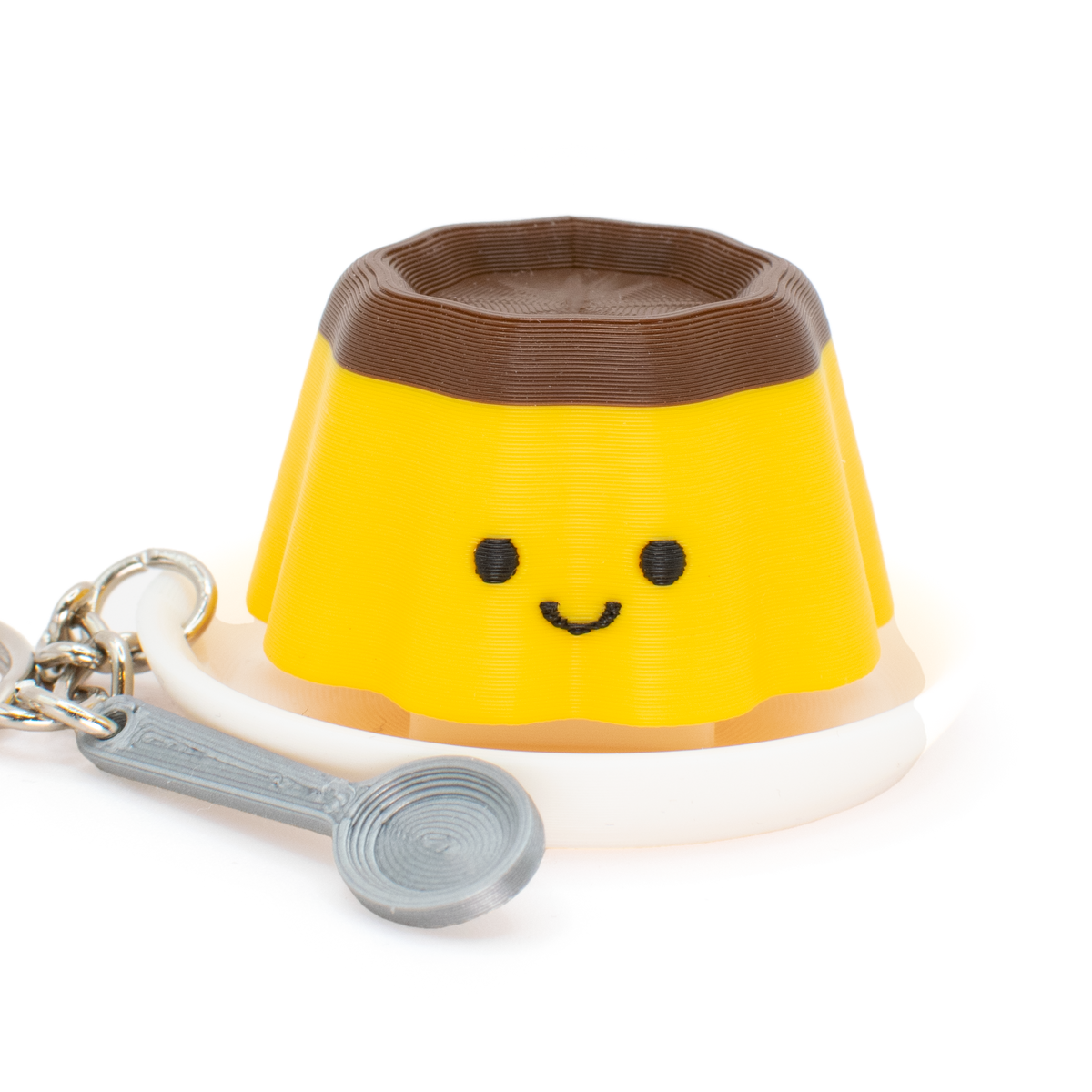 A yellow and brown clicker keychain shaped like a smiling pudding, featuring with a white plate and a silver spoon charm attached with a metal keyring, shown on a plain white background.