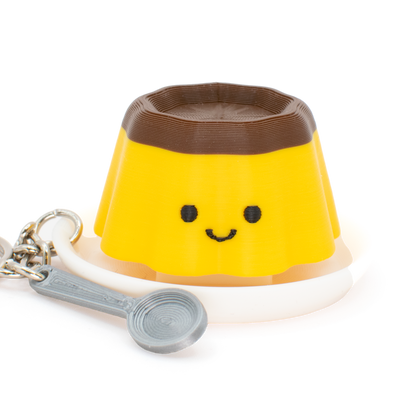 A yellow and brown clicker keychain shaped like a smiling pudding, featuring with a white plate and a silver spoon charm attached with a metal keyring, shown on a plain white background.