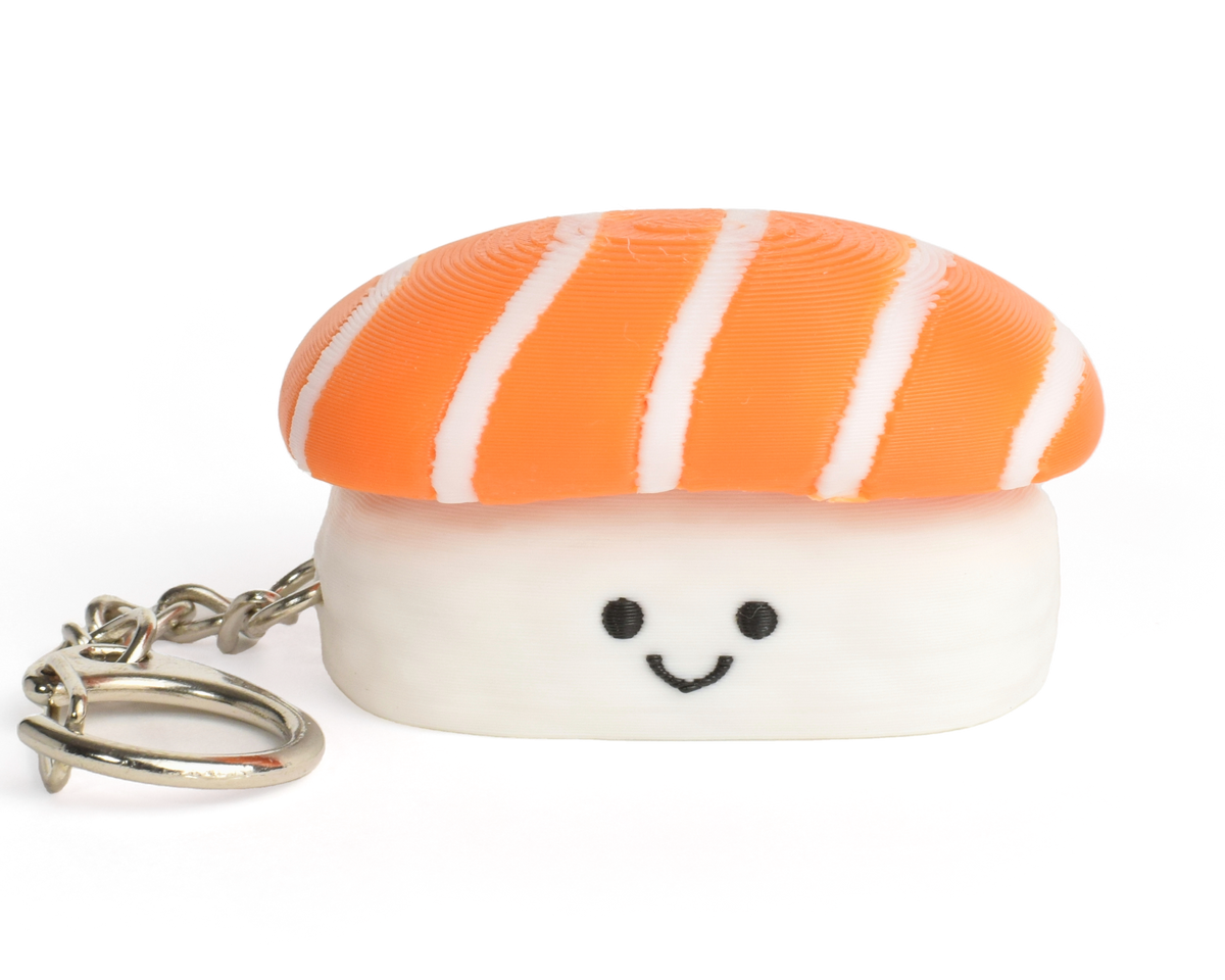 A small sushi-shaped clicker keychain with a smiling face, featuring a salmon top and a white rice base, attached to a silver metal key ring on a plain white background.