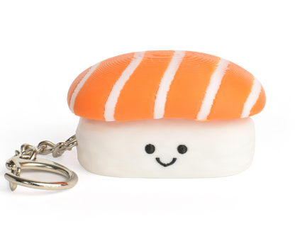 A small sushi-shaped clicker keychain with a smiling face, featuring a salmon top and a white rice base, attached to a silver metal key ring on a plain white background.