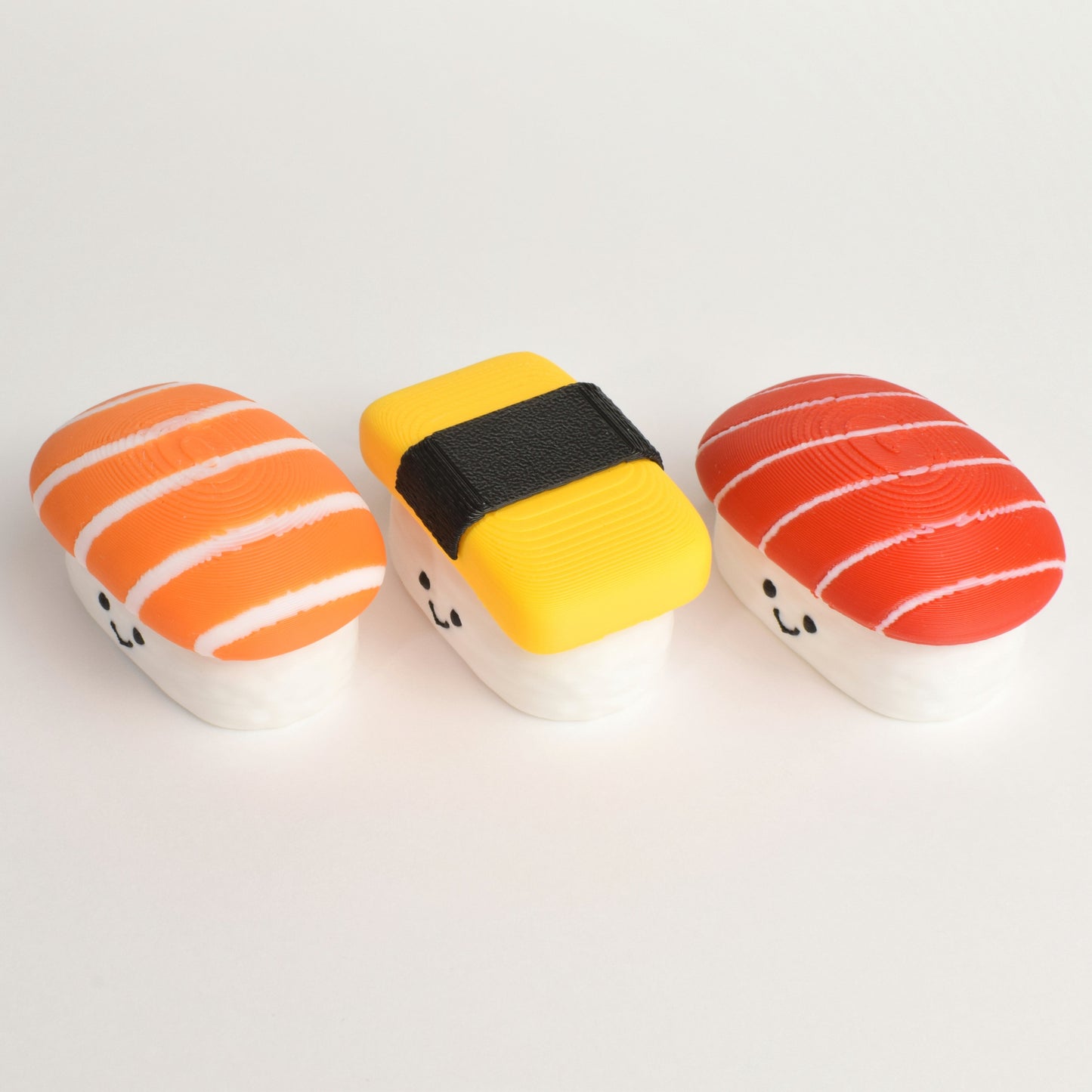 Three small sushi-shaped clickers arranged on a plain white background: they are salmon nigiri sushi, tuna nigiri sushi, and tamago (egg) nigiri sushi