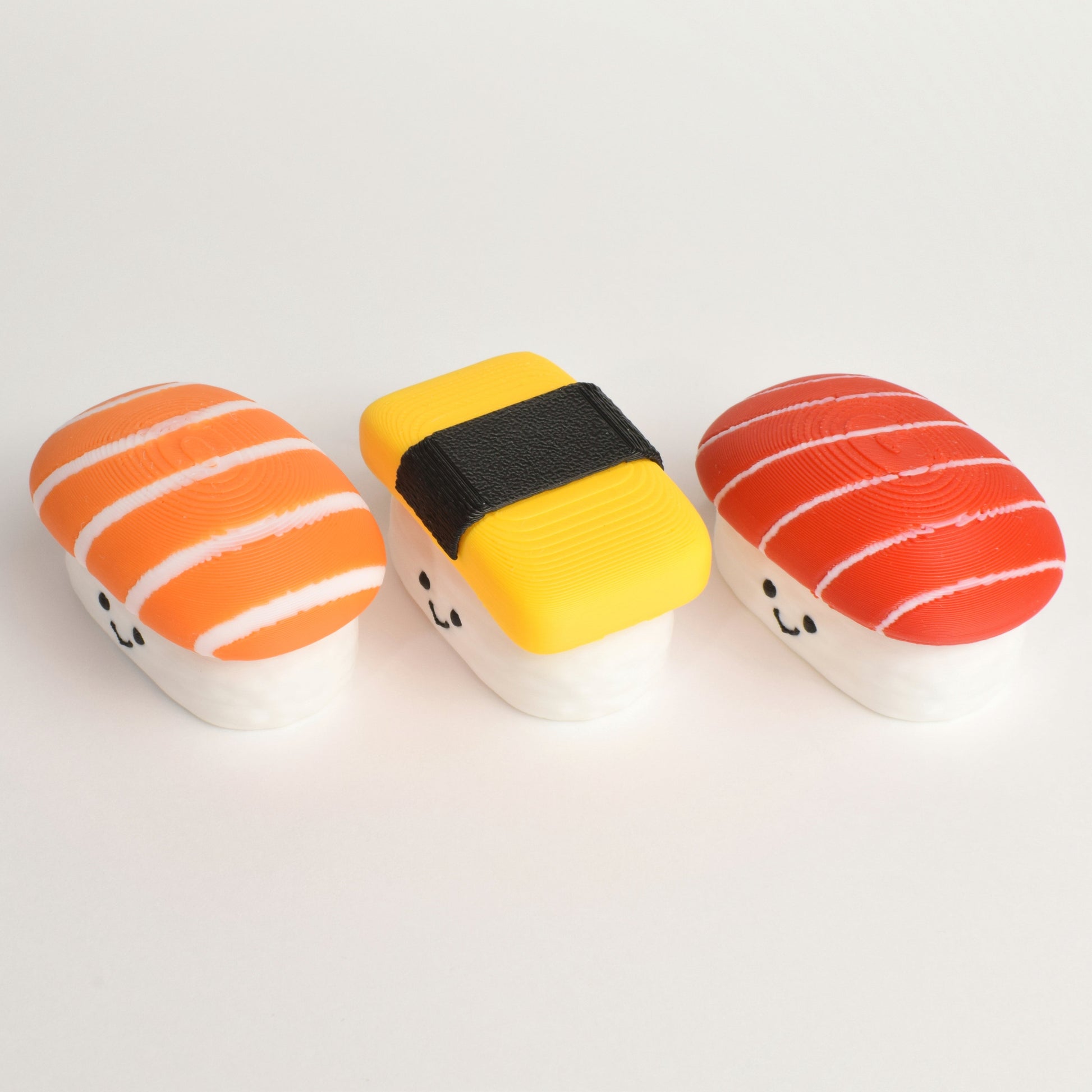 Three small sushi-shaped clickers arranged on a plain white background: they are salmon nigiri sushi, tuna nigiri sushi, and tamago (egg) nigiri sushi