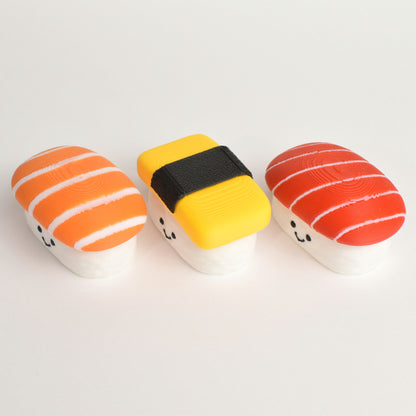 Three small sushi-shaped clickers arranged on a plain white background: they are salmon nigiri sushi, tuna nigiri sushi, and tamago (egg) nigiri sushi