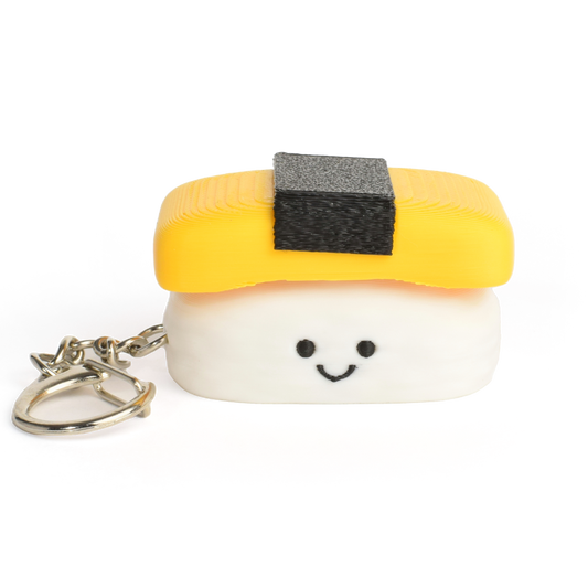 A small sushi-shaped clicker keychain with a smiling face, featuring a yellow egg top and a white rice base, attached to a silver metal key ring on a plain white background.