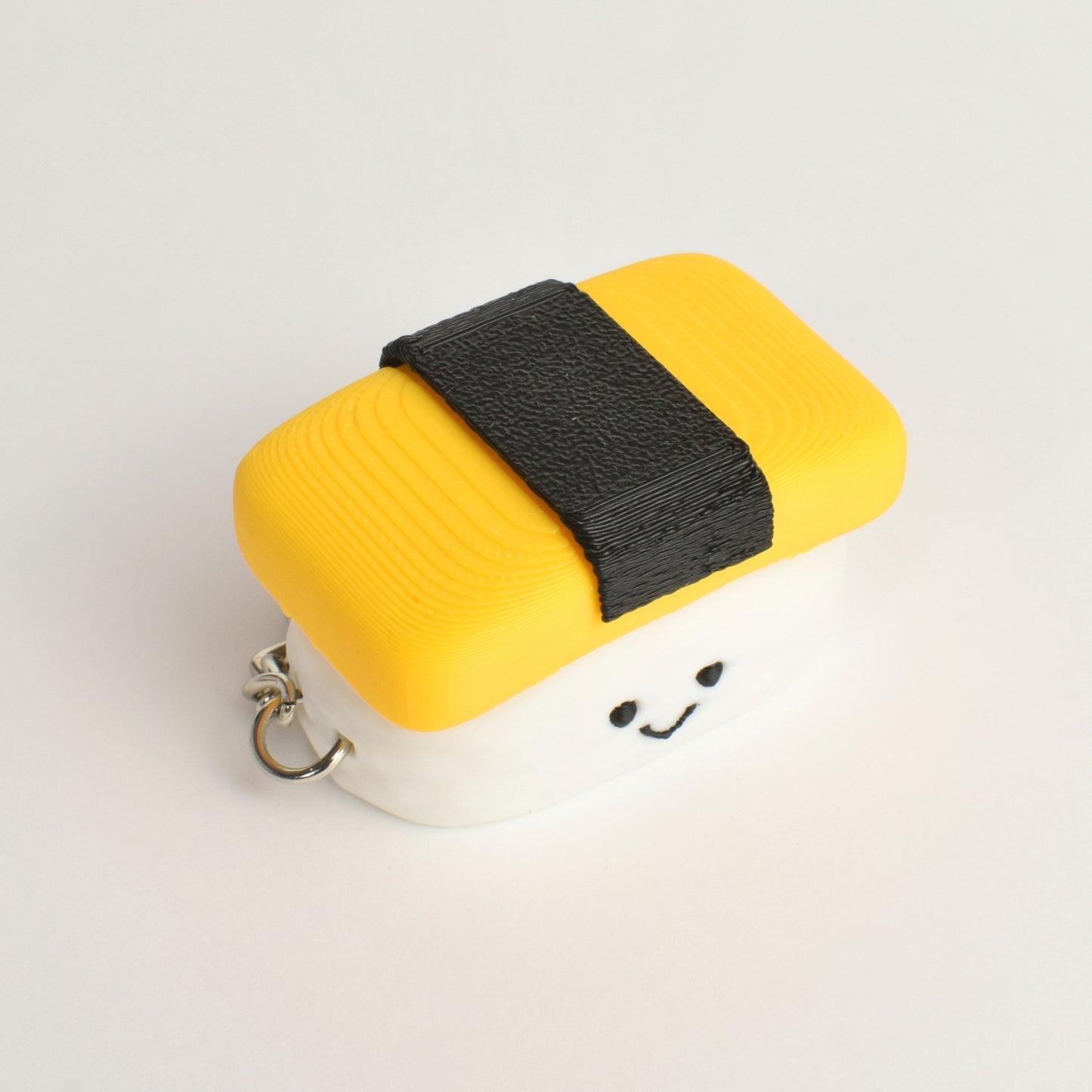 A small novelty keychain shaped like a piece of sushi, featuring a yellow egg top with seaweed and a white base with a cute smiling face and a metal keyring.