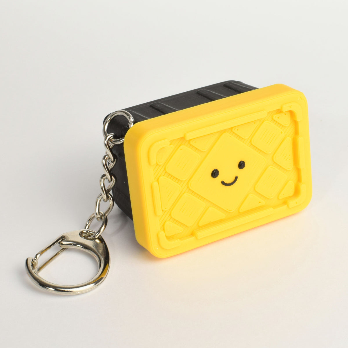 A small keychain shaped like a tote storage bin with a smiling face, attached to a silver metal key ring, photographed on a clean white background.
