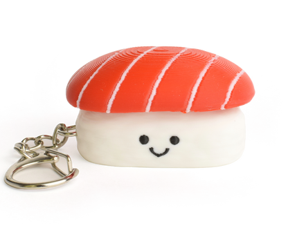 A small sushi-shaped clicker keychain with a smiling face, featuring a tuna top and a white rice base, attached to a silver metal key ring on a plain white background.