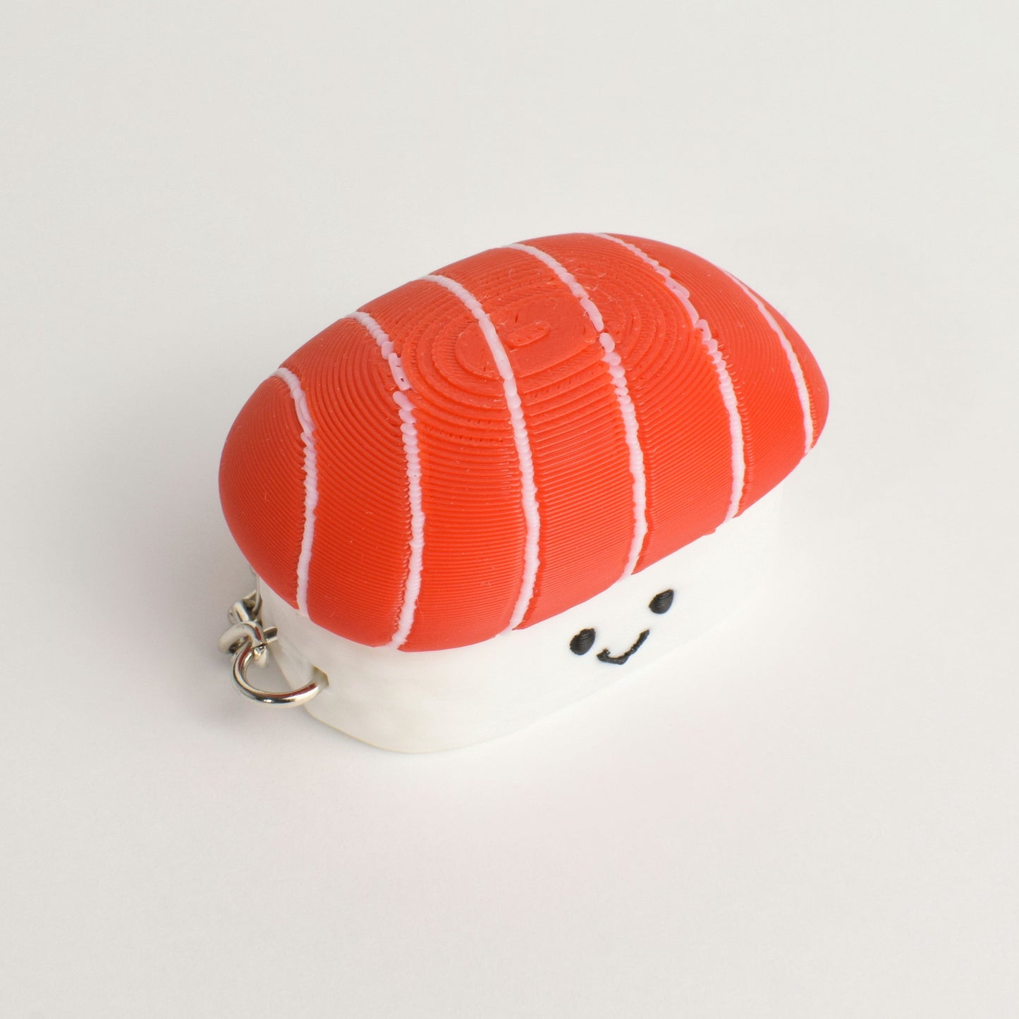 top view of a small sushi-shaped clicker keychain with a smiling face, featuring a tuna top and a white rice base, attached to a silver metal key ring on a plain white background.