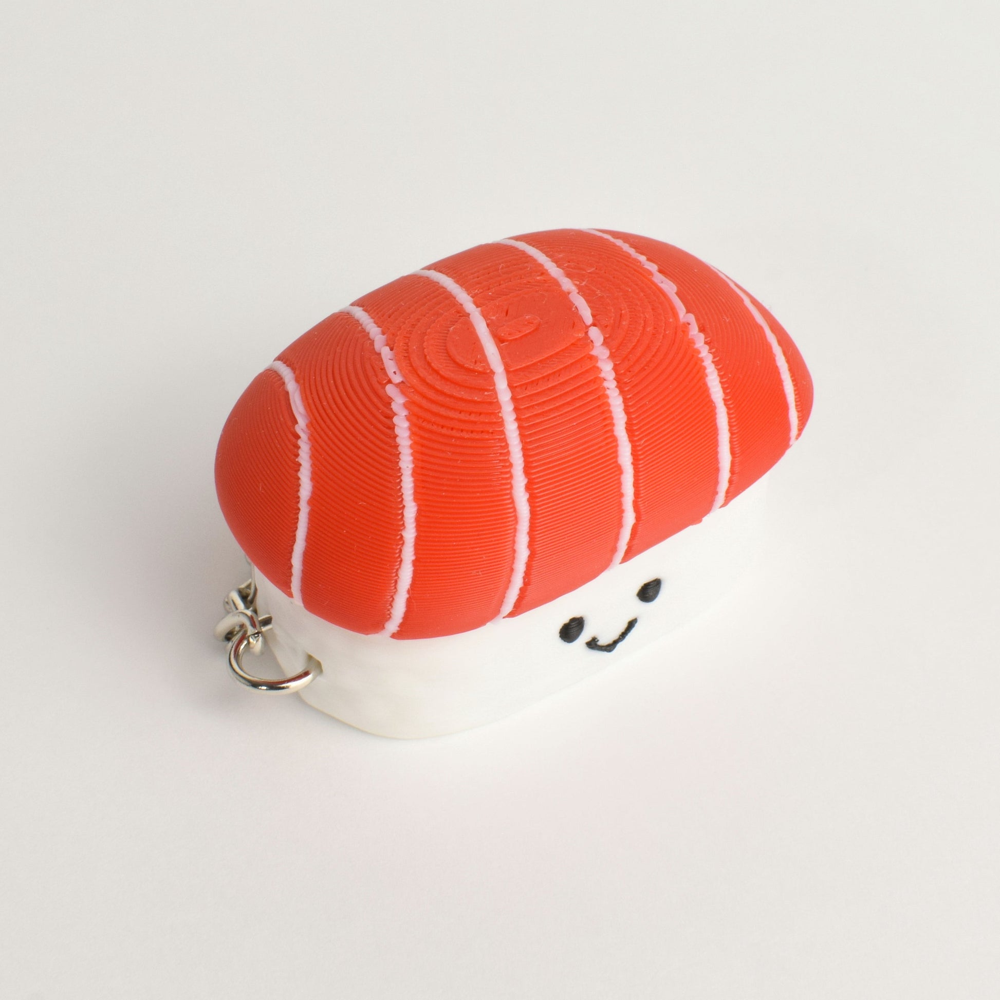 top view of a small sushi-shaped clicker keychain with a smiling face, featuring a tuna top and a white rice base, attached to a silver metal key ring on a plain white background.